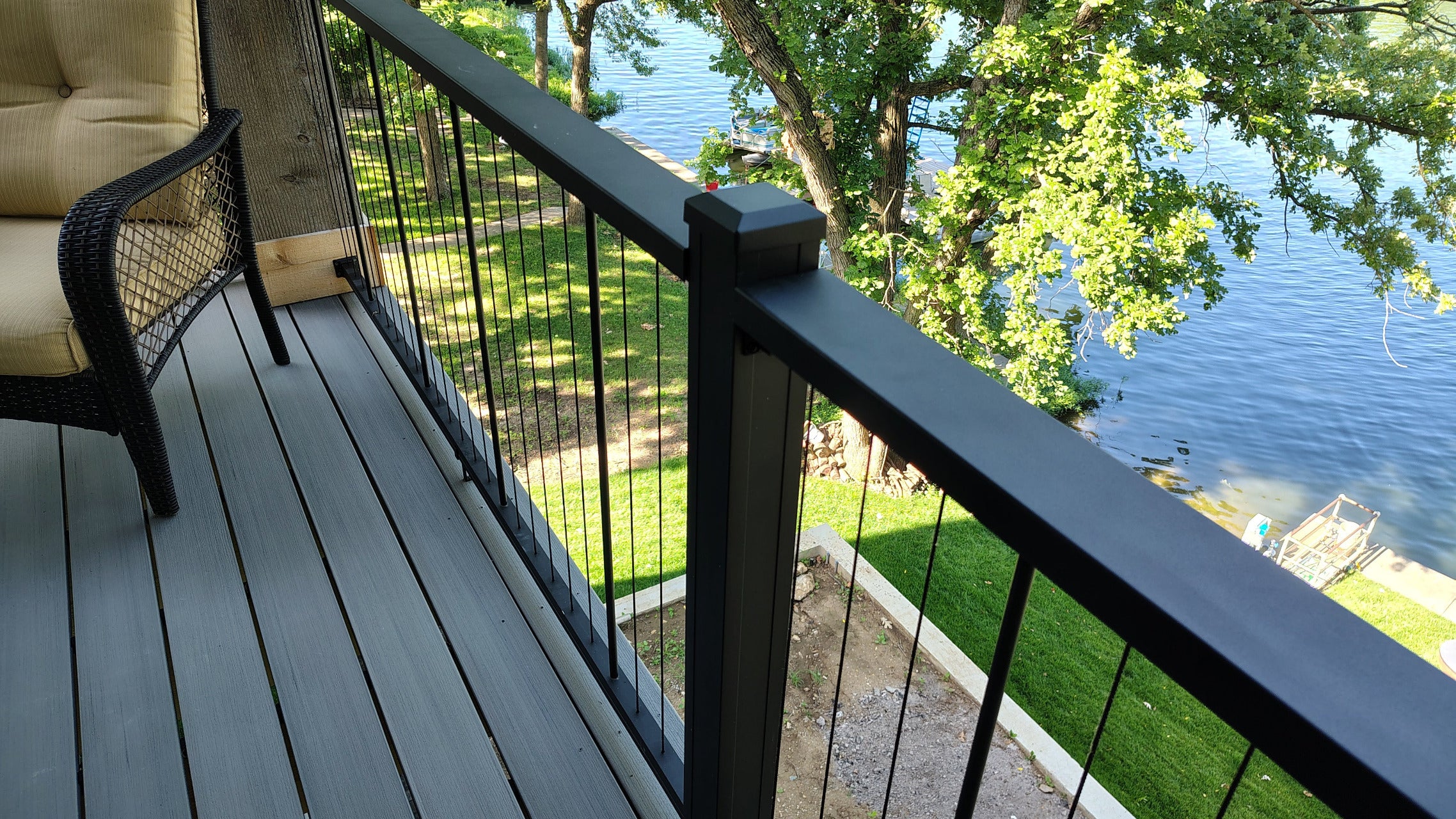 A close-up view of modern vertical cable deck railing system Chesapeake Vertical Cable by KeyLink with dark gray posts and cables, installed on a gray composite deck overlooking a green lawn and a lake with trees.
