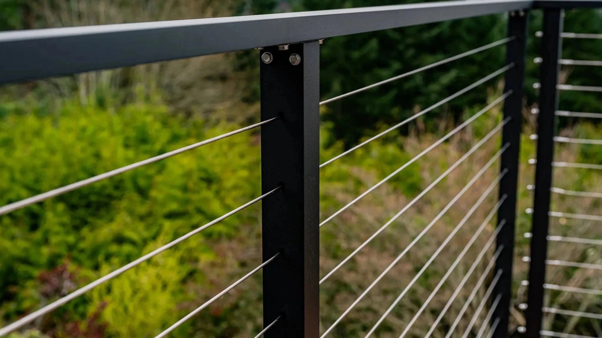 Close-up of a Maisy ultra-modern black metal cable railing on a wooden deck with a blurred green garden background.