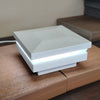 A modern square LED light Cap from magena star - white texture ornamental lighted post cap box with a textured light gray top and white sides, glowing with a bright white light band, resting on a wooden surface.