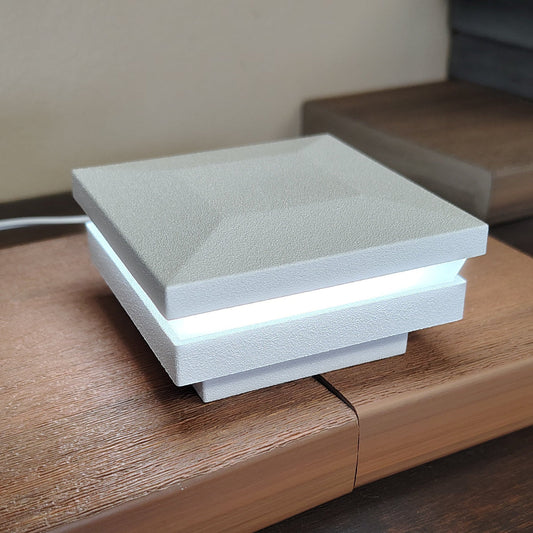 A modern square LED light Cap from magena star - white texture ornamental lighted post cap box with a textured light gray top and white sides, glowing with a bright white light band, resting on a wooden surface.