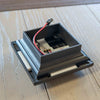 A close-up of a black 2.5" lighted post cap with lighting component and a red wire attached, resting on a light-colored wooden surface.