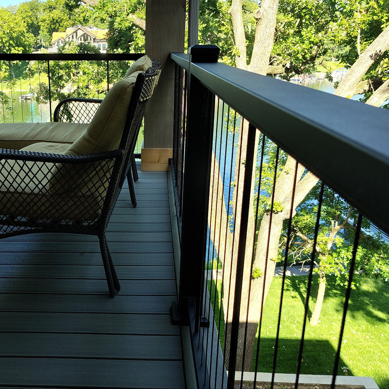 KeyLink Black Cable Vertical Deck Rail  Kits on a dark gray composite deck with outdoor furniture, including a wicker armchair with a beige cushion, a matching coffee table, and a chaise lounge, overlooking a green lawn and trees from a balcony.