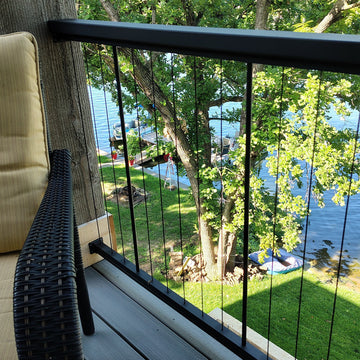 KeyLink Chesapeake Level Rail Kit with Vertical Black Cable Rail with Top Cap (top cap sold separately), with a view from the composite wood deck overlooking a green lawn, trees, and a body of water, with a black wicker chair and beige cushion visible in the foreground.