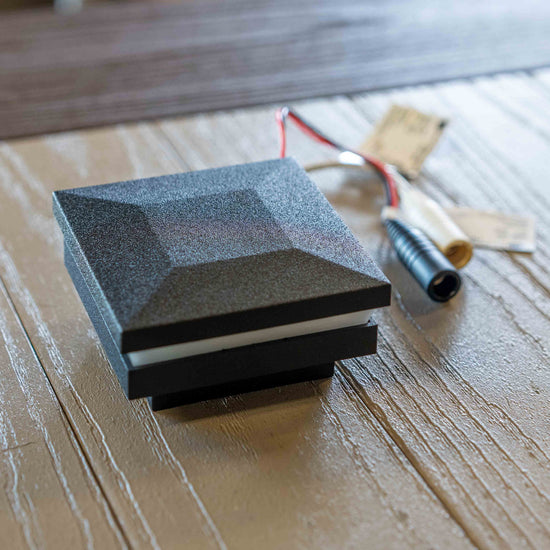 A square black LED deck cap light rail fixture with a textured geometric top, shown on a wooden surface next to its mounting hardware.