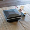 A square black LED deck cap light rail fixture with a textured geometric top, shown on a wooden surface next to its mounting hardware.