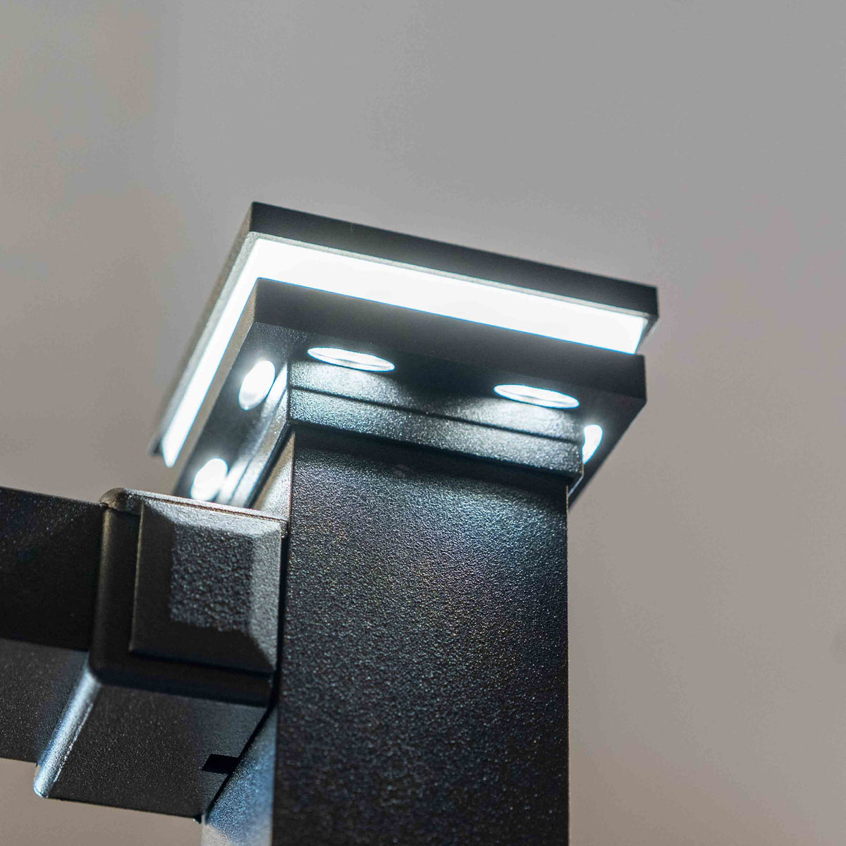 Close-up of a modern black LED light fixture mounted on a wall, with the top panel illuminated and multiple light elements visible against a plain light background.