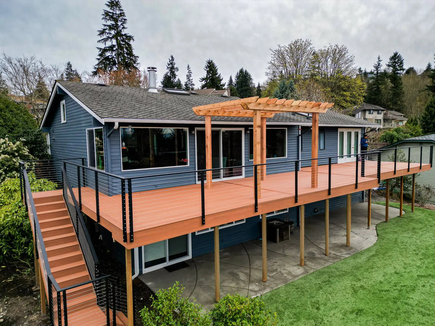 A two-story house with dark blue siding and a large orange-brown wooden deck with a matching pergola, outdoor stairs, black railings, and a green lawn under an overcast sky.