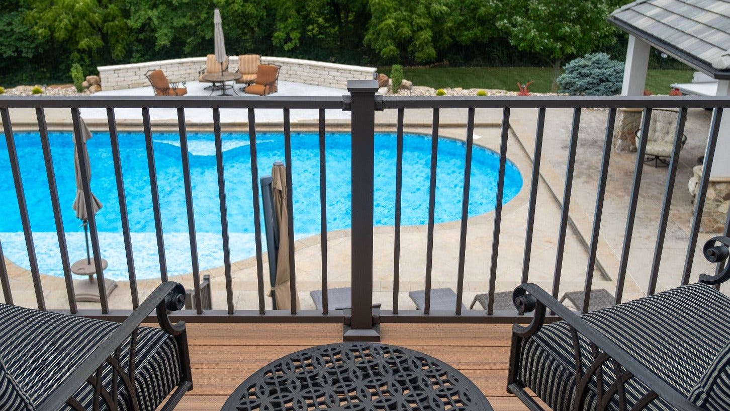 Outdoor deck with pool, chairs, and table, surrounded by greenery with aluminum deck rail and composite decking