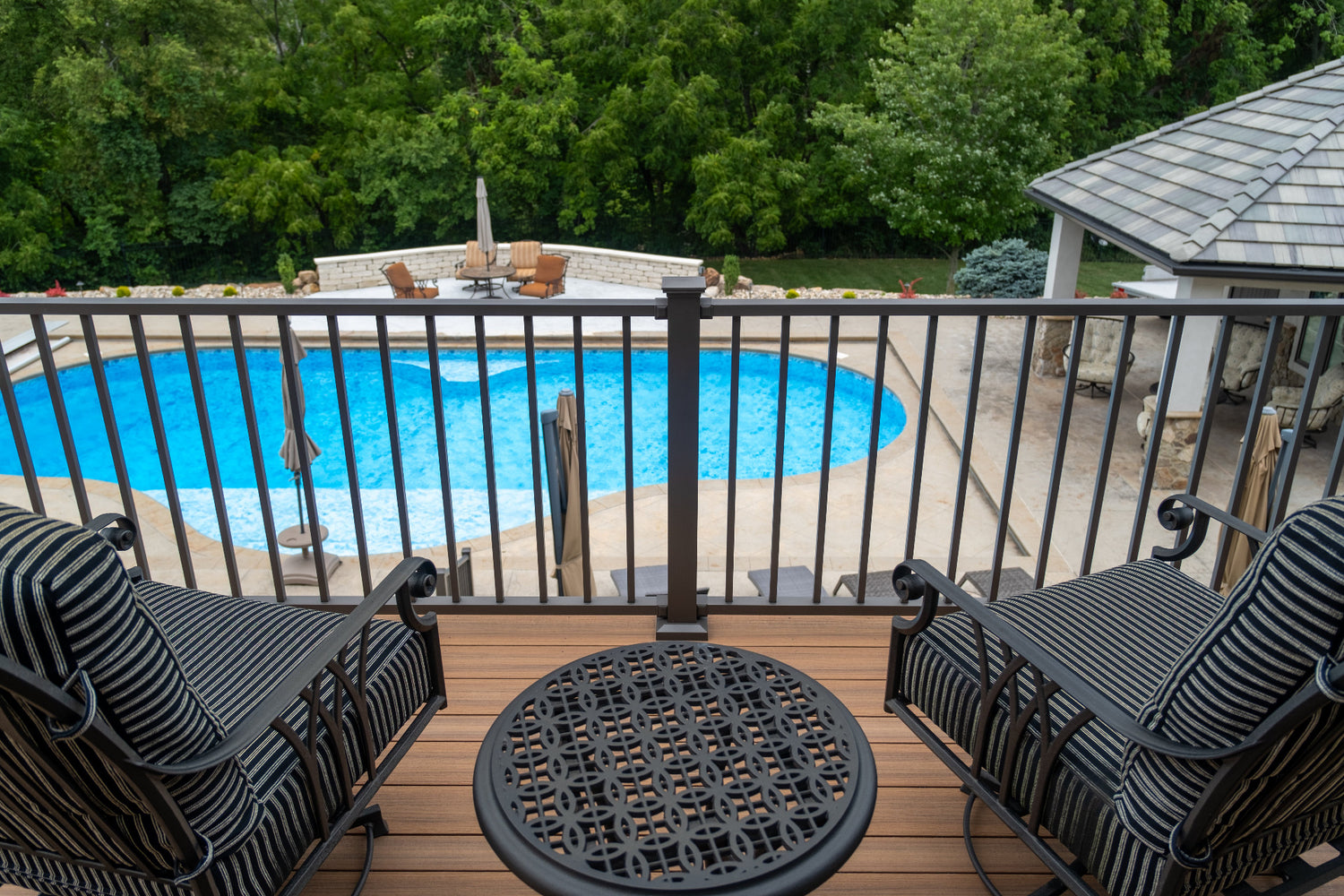 Outdoor deck with pool, chairs, and table, surrounded by greenery with aluminum deck rail and composite decking