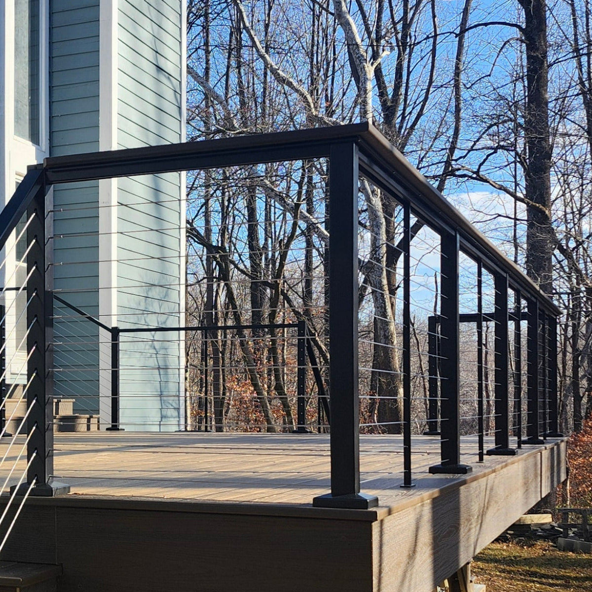 Black Feeney Modern metal cable railing system installed on a wooden deck outdoors, with bare trees and a house visible in the background.