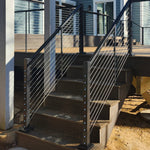Feeney DesignRailKits Stair Rail Outdoor staircase with black metal 250 stair rail kits and a wooden deck platform attached to a blue house, shown in a backyard setting with bare trees and dirt ground.