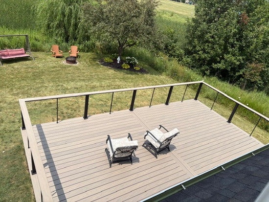 A high-angle view of a wooden deck with a light brown composite finish and black metal railings, furnished with two patio chairs. The deck is situated on a grassy backyard with trees, lawn, and outdoor seating visible in the background.