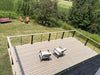 A high-angle view of a wooden deck with a light brown composite finish and black metal railings, furnished with two patio chairs. The deck is situated on a grassy backyard with trees, lawn, and outdoor seating visible in the background.