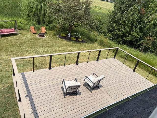 A high-angle view of a wooden deck with a light brown composite finish and black metal railings, furnished with two patio chairs. The deck is situated on a grassy backyard with trees, lawn, and outdoor seating visible in the background.