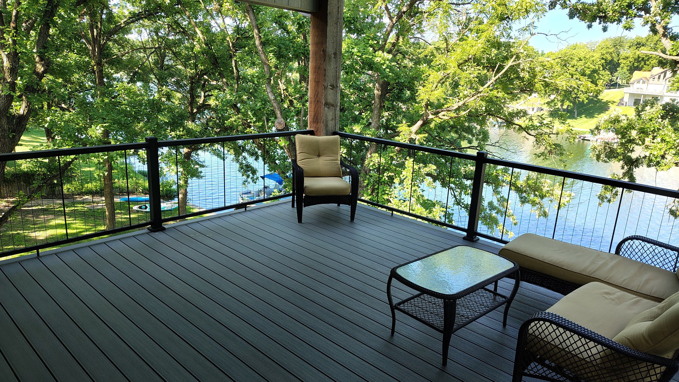 KeyLink Chesapeake Vertical Cable with a dark gray composite deck with outdoor furniture, including a chair, a loveseat with beige cushions, and a small glass-top coffee table, overlooking a lake and green trees.