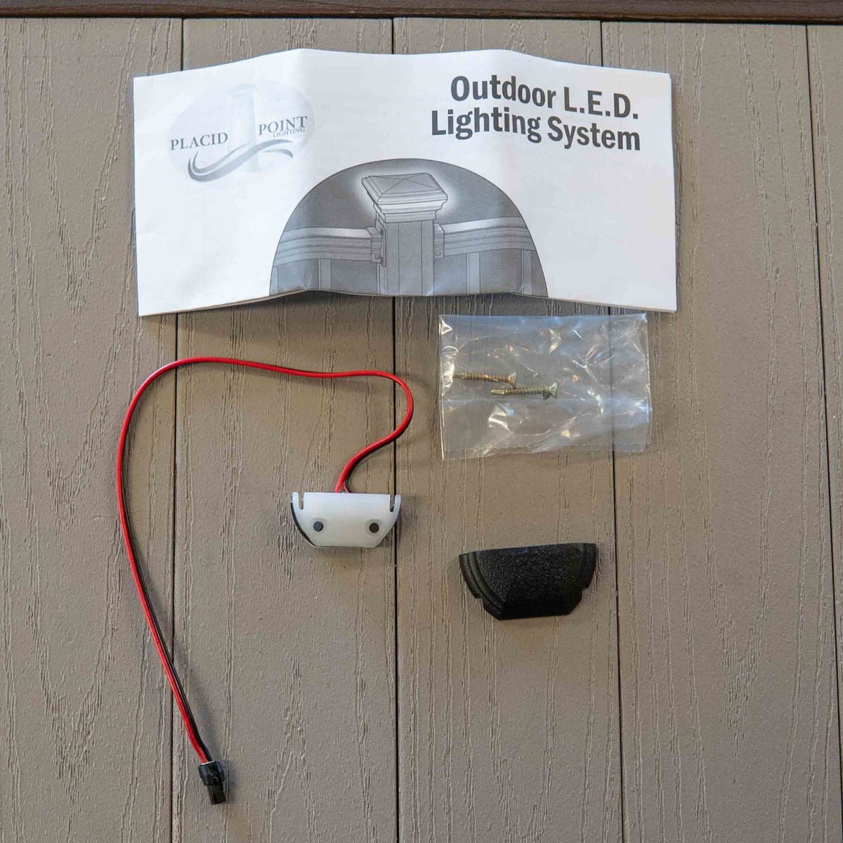 Outdoor LED lighting system component KeyLink Post Accent Light for Posts and Stairs laid out on a porch deck panel background, including a white printed instruction sheet, a red-and-black wire with a small white component, a black cap, and a clear plastic bag of small metal parts.